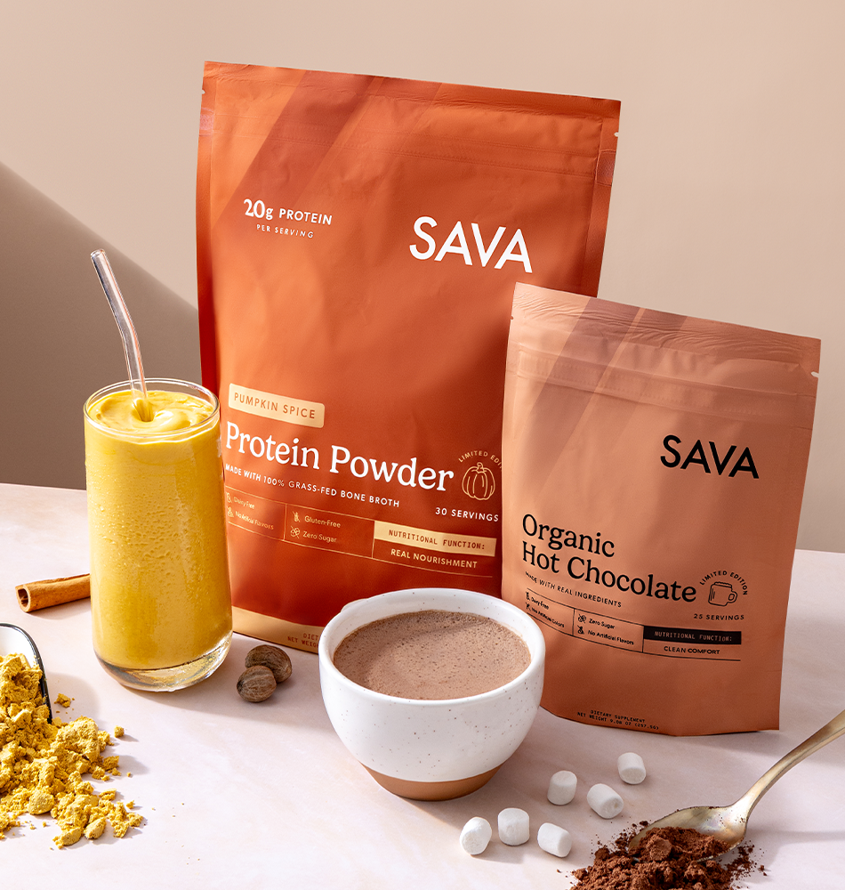 Two SAVA branded pouches of protein powder stand on a tabletop, one in orange packaging labeled Protein Powder and one in brown packaging labeled Organic Hot Chocolate. A glass of yellow drink, a bowl of hot chocolate, scattered cocoa powder, marshmallows, and a spoon are arranged around them on a light beige surface.
