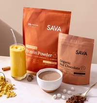 Two SAVA branded pouches of protein powder stand on a tabletop, one in orange packaging labeled Protein Powder and one in brown packaging labeled Organic Hot Chocolate. A glass of yellow drink, a bowl of hot chocolate, scattered cocoa powder, marshmallows, and a spoon are arranged around them on a light beige surface.